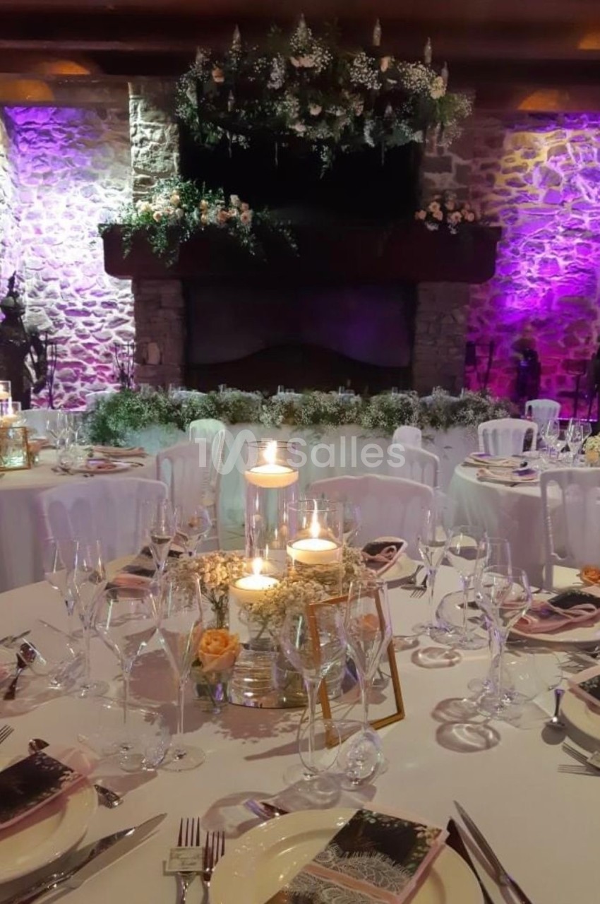 Salle de réception décorée pour un mariage, avec tables dressées, bougies et éclairage violet sur des murs en pierre.