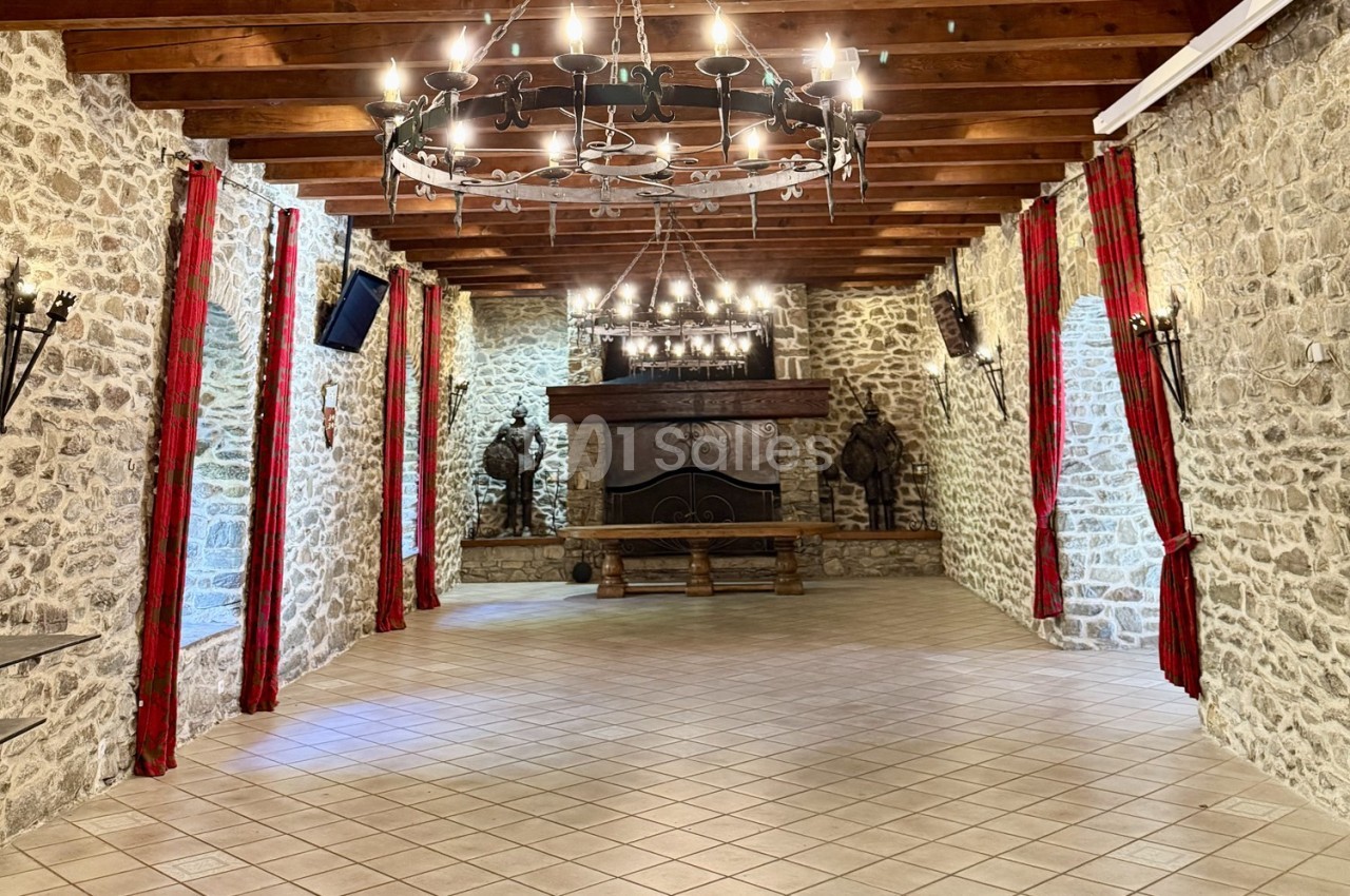 Salle en pierre avec un plafond en poutres apparentes, lustres suspendus, cheminée et rideaux rouges encadrant les murs.