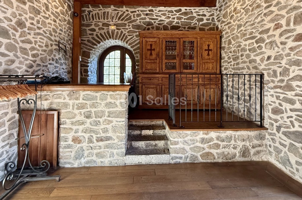 Intérieur en pierre avec un escalier menant à une mezzanine, meuble en bois massif et porte-fenêtre voûtée.