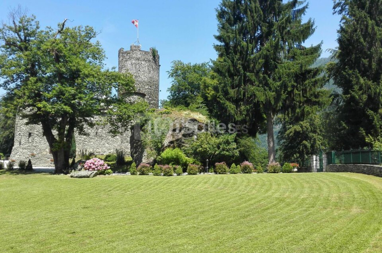 Château médiéval en pierre entouré de verdure, avec un drapeau au sommet et une pelouse bien entretenue au premier plan.