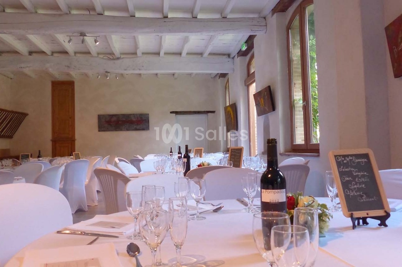 Salle de réception lumineuse avec tables dressées, nappes blanches, verres, bouteilles de vin et ardoise décorative.