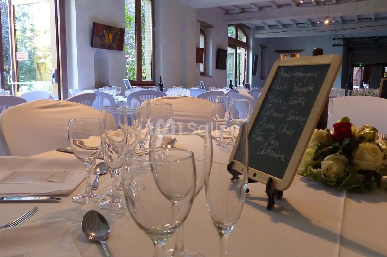 Salle de réception lumineuse avec tables dressées, nappes blanches, verres, couverts et ardoise de menu.