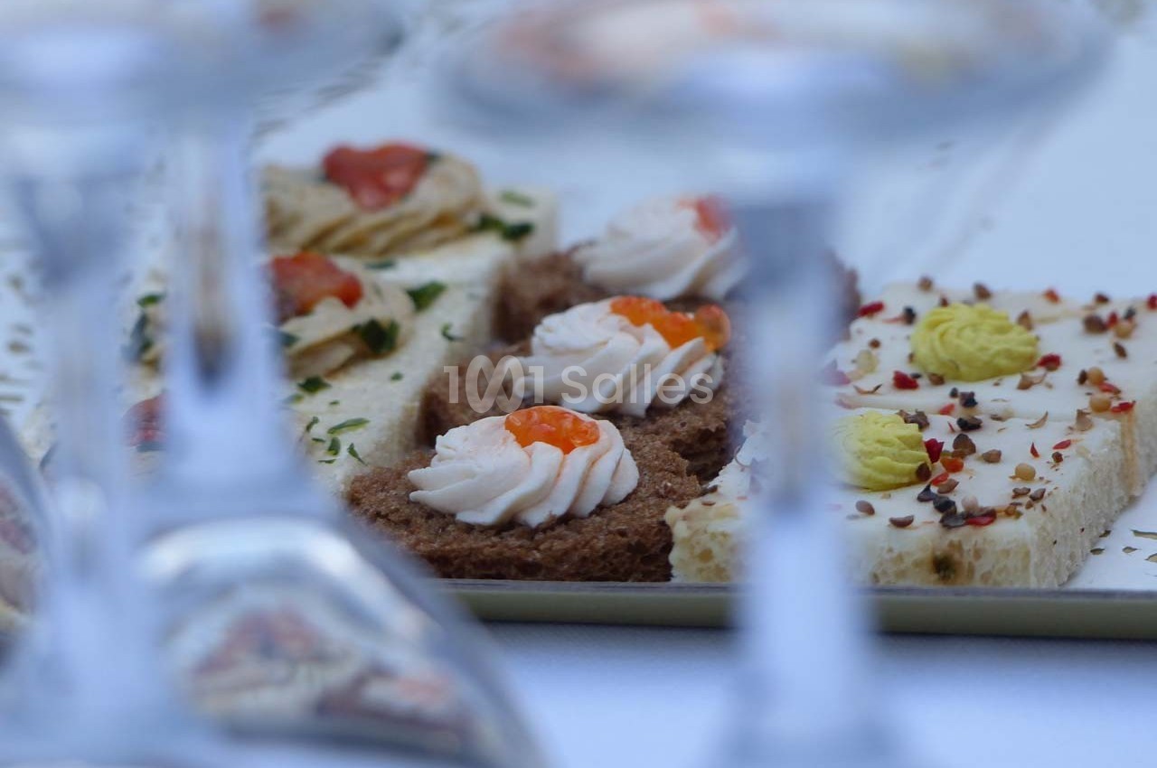 Assortiment de canapés garnis de mousse et de condiments, présenté sur un plateau derrière des verres floutés.