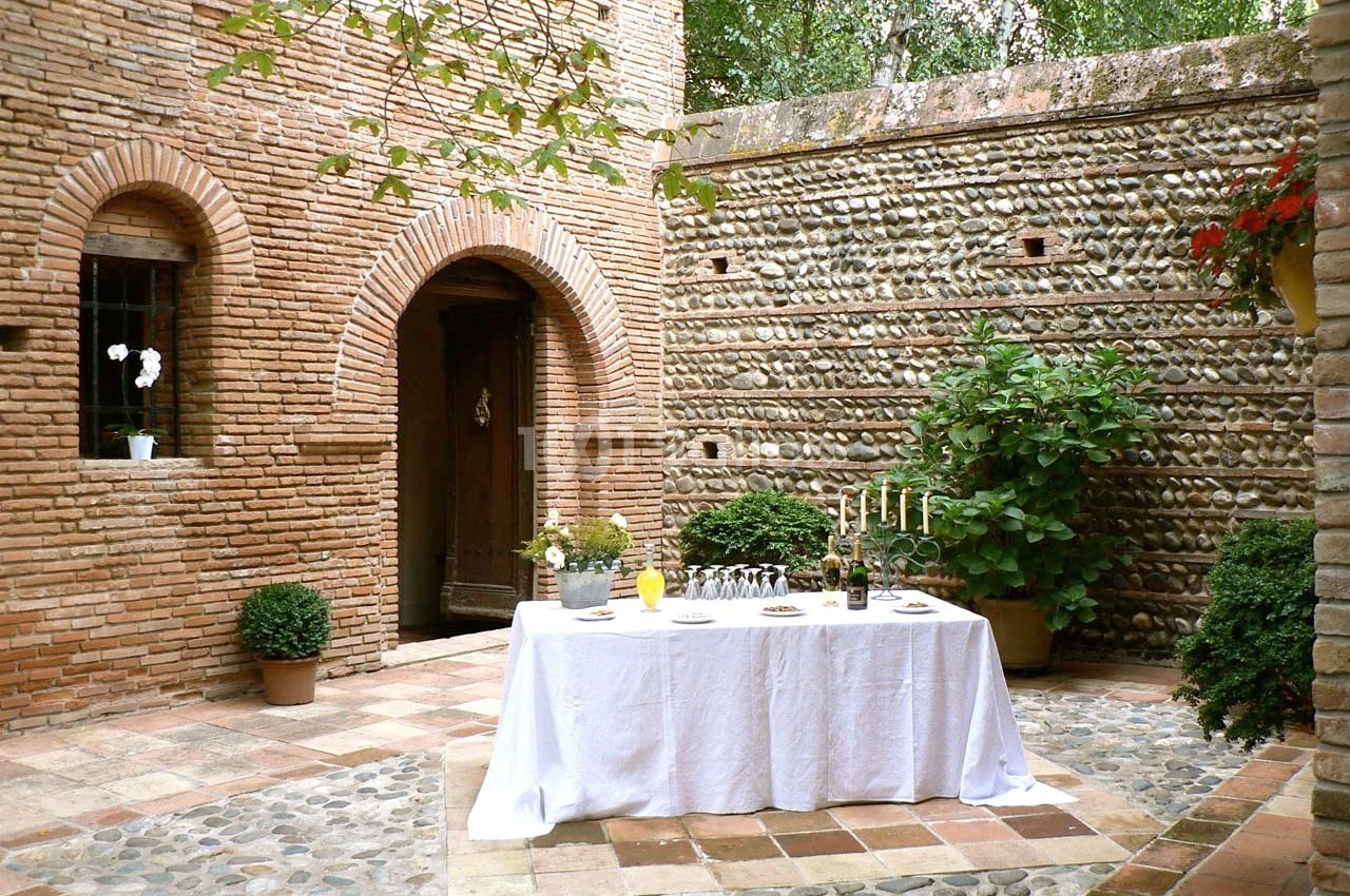 Table dressée avec nappes blanches dans une cour en briques et galets, entourée de plantes et d'une porte en bois.