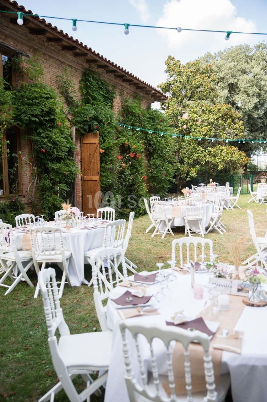 Tables rondes décorées pour un événement en plein air, disposées dans un jardin avec une façade en briques et des guirlandes…