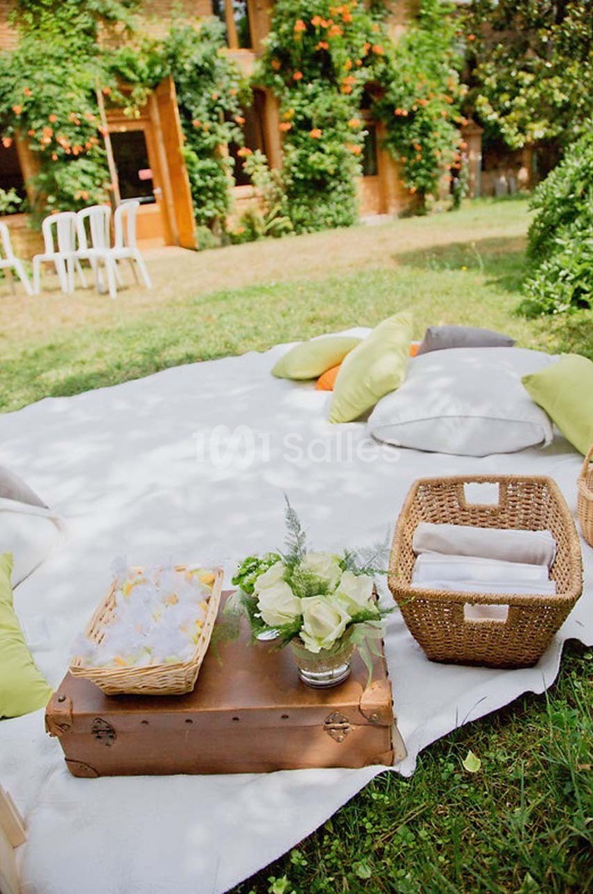 Tapis blanc sur pelouse avec coussins colorés, paniers en osier, fleurs et table basse en bois dans un jardin verdoyant.