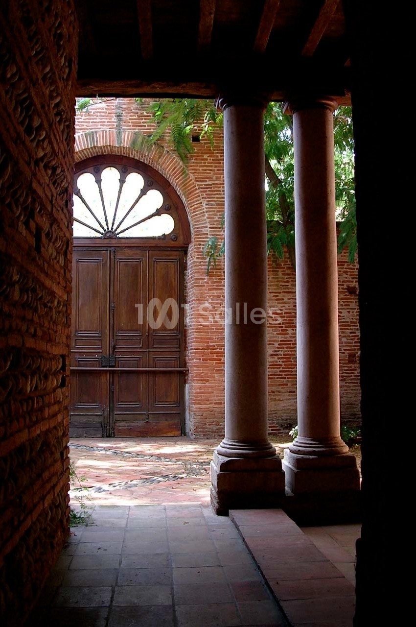 Vue d'une porte en bois encadrée par des colonnes et un mur de briques, éclairée par la lumière extérieure.