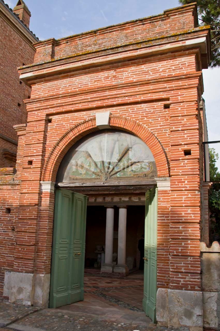 Entrée en briques rouges avec une arche en pierre, portes vertes ouvertes et un intérieur visible avec des colonnes.