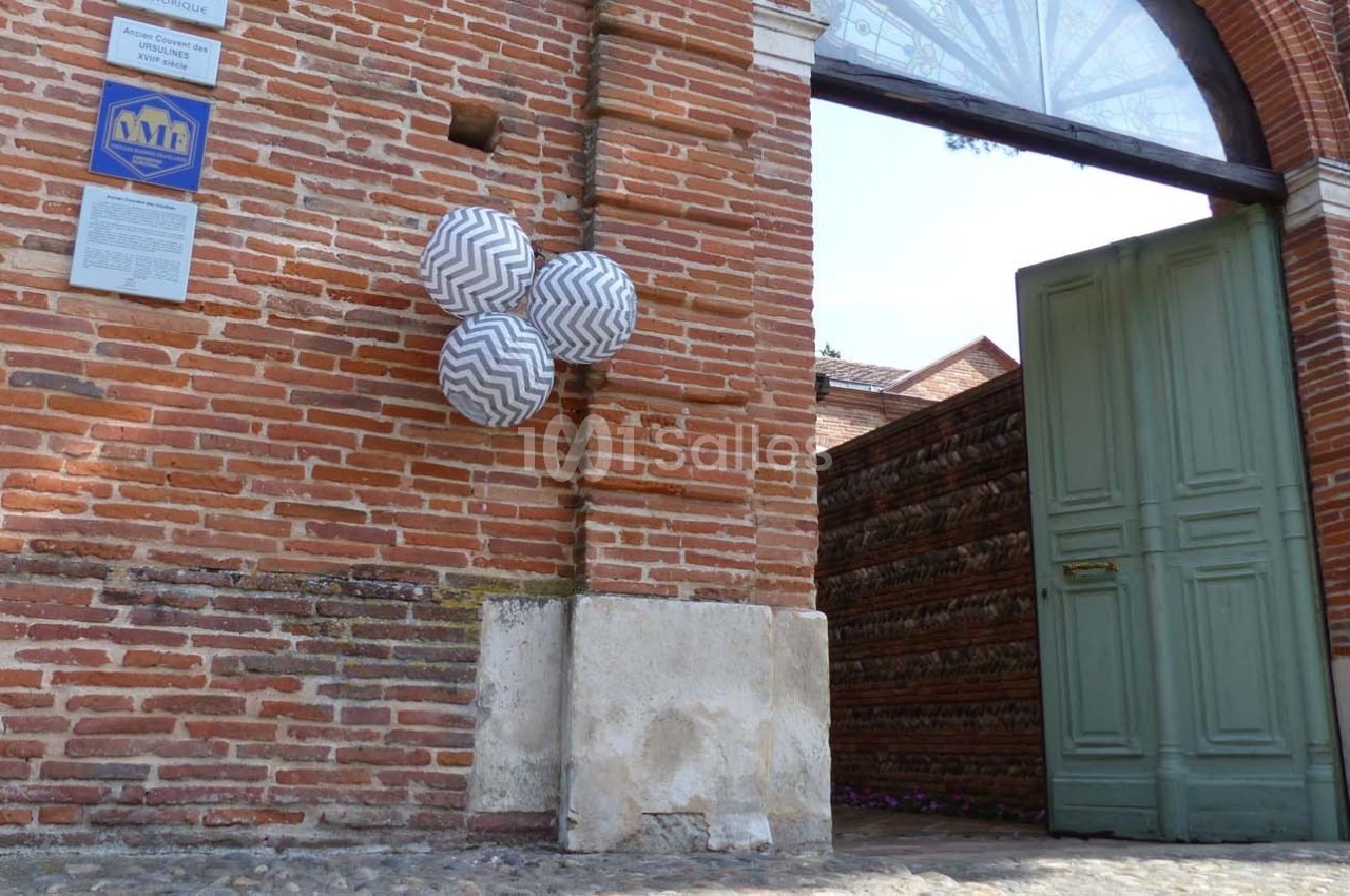 Entrée en briques rouges avec une porte verte ouverte et des lanternes suspendues à motifs zigzag.