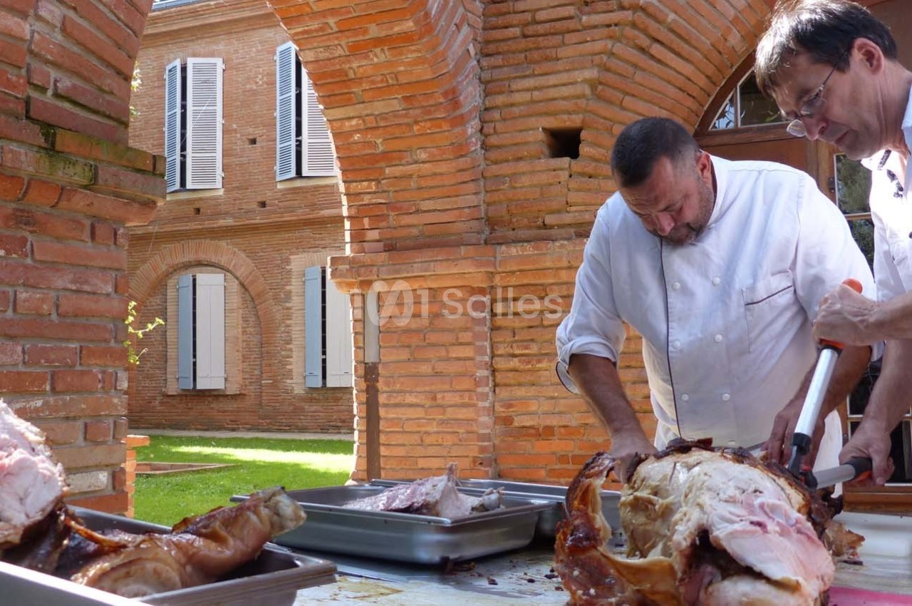 Deux chefs découpent un cochon rôti sur une table en extérieur, près d'une maison en briques rouges.
