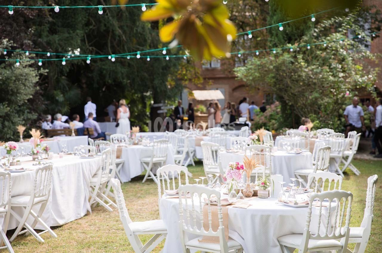 Tables décorées pour un événement en plein air, entourées de verdure et éclairées par des guirlandes lumineuses.