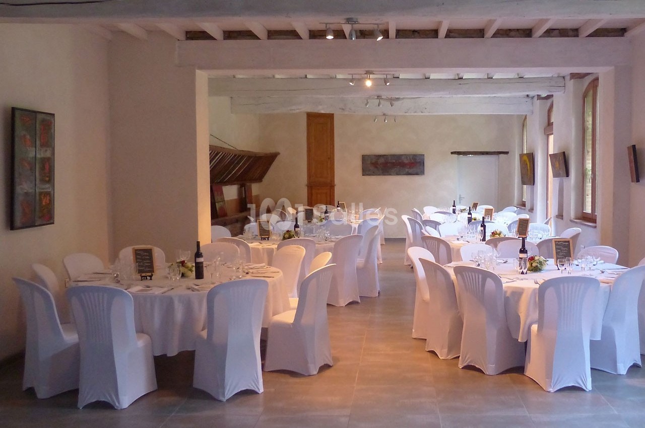 Salle de réception lumineuse avec tables rondes dressées, nappes blanches et chaises couvertes, prête pour un événement.