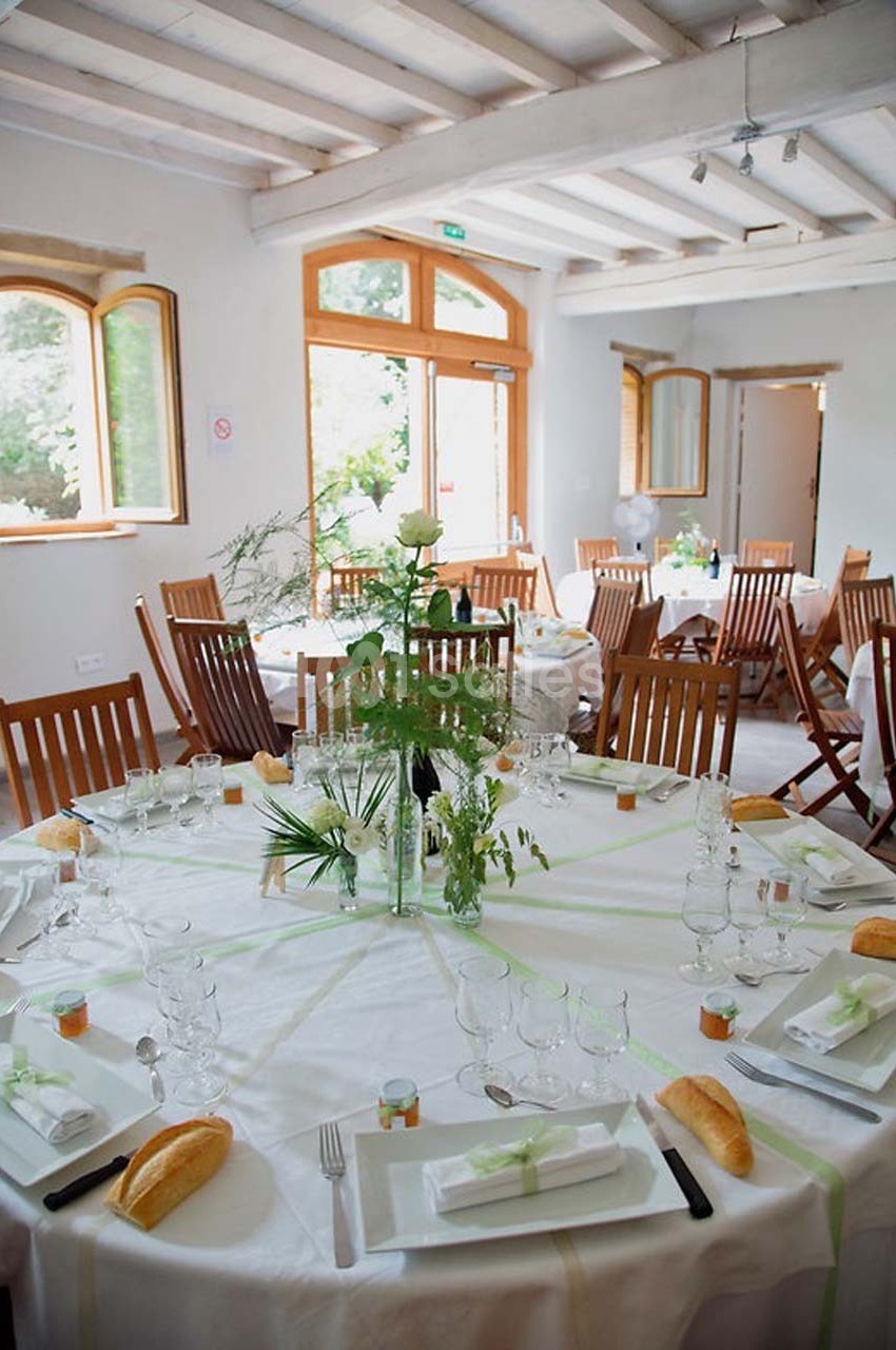 Salle lumineuse avec tables rondes dressées pour un repas, décorées de nappes blanches, plantes et vaisselle élégante.