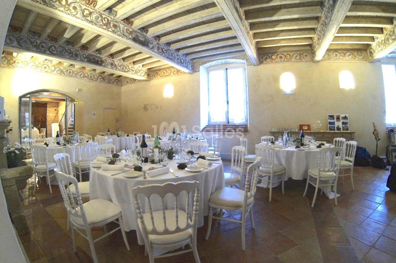 Salle de réception avec tables rondes dressées, chaises blanches et plafond en bois sculpté.