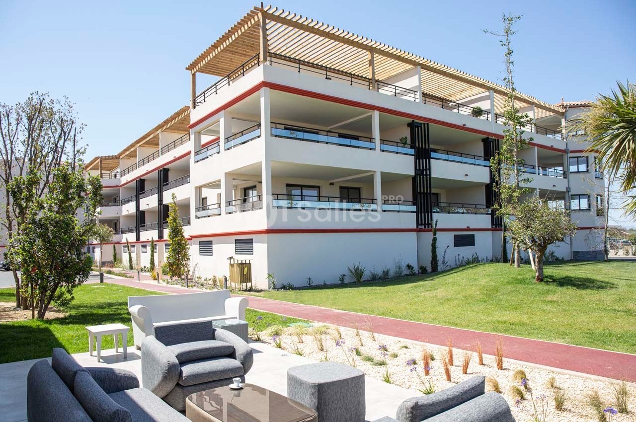 Résidence moderne à trois étages avec balcons, entourée d'espaces verts et d'un coin salon extérieur aménagé.