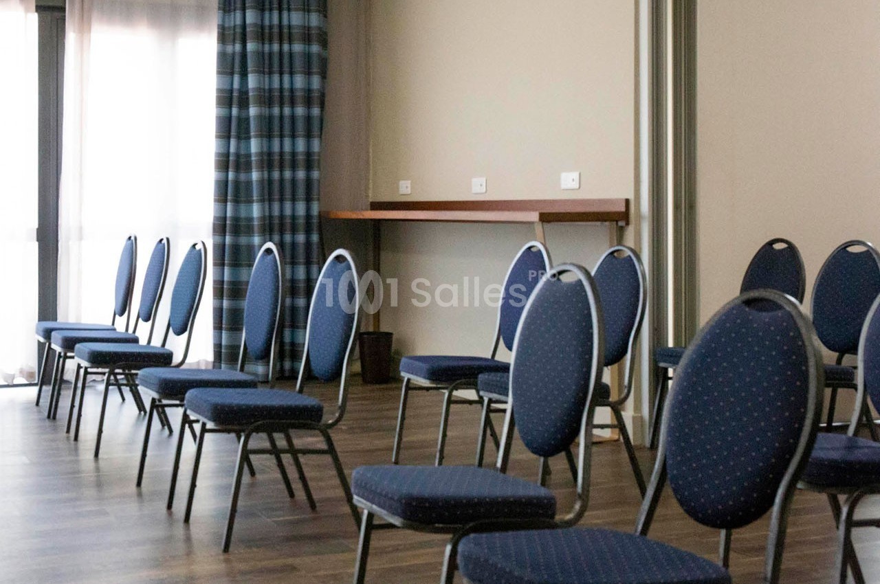 Chaises bleues disposées en rangées dans une salle lumineuse avec parquet et rideaux à carreaux.