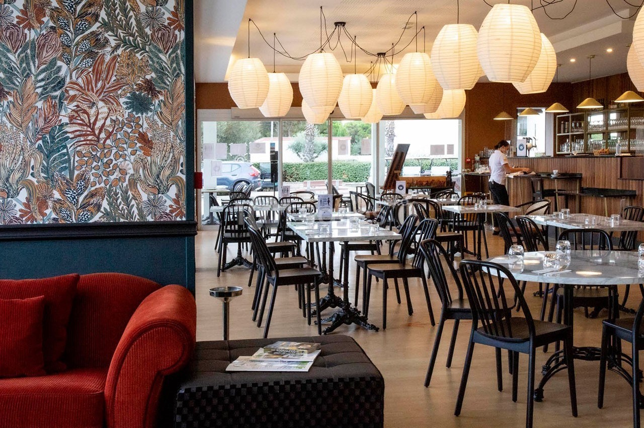 Salle de restaurant lumineuse avec tables en bois, chaises noires, lampes suspendues et un mur décoré de motifs végétaux.