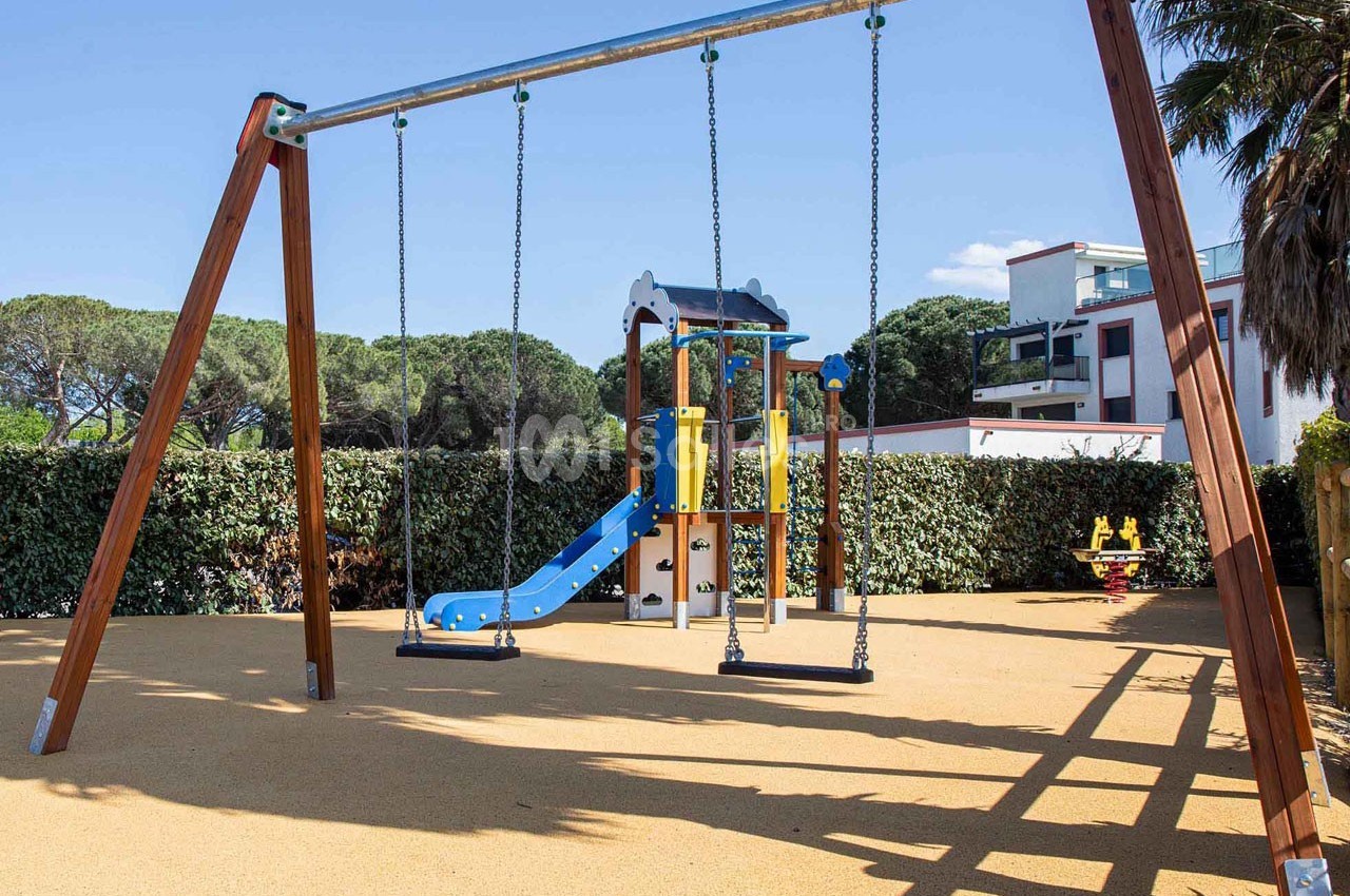 Structure de jeux pour enfants avec toboggan et balançoires, installée sur un sol sablé entouré de végétation.