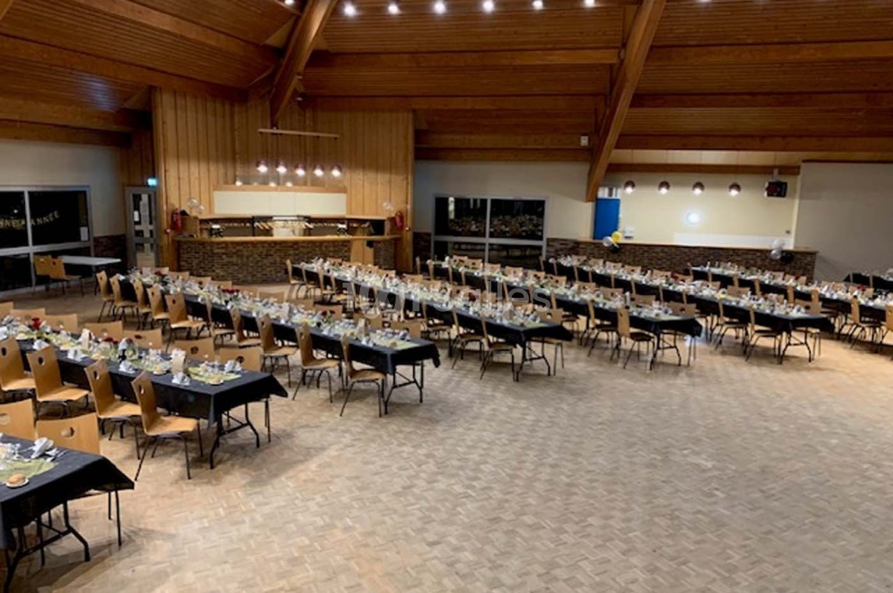 Salle spacieuse avec des tables rectangulaires dressées pour un événement, éclairée par des lumières suspendues.