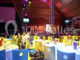 Salle de réception avec des tables rondes dressées, éclairage coloré et écrans suspendus.