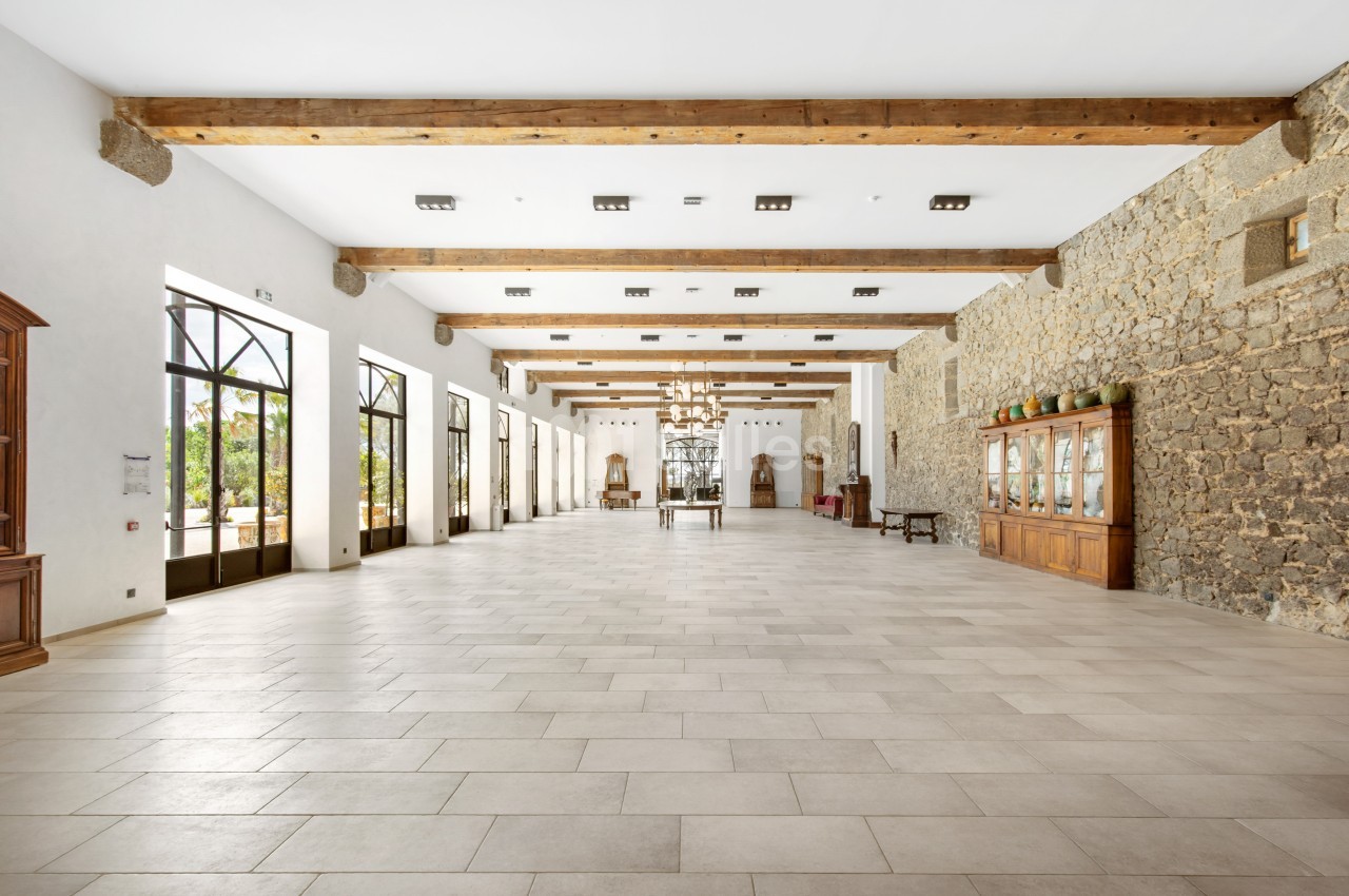 Grande salle lumineuse avec murs en pierre, poutres apparentes, carrelage clair et grandes fenêtres donnant sur l'extérieur.