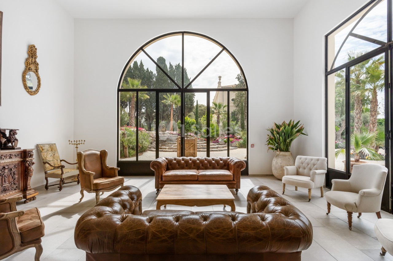 Salon lumineux avec canapés en cuir, fauteuils classiques et vue sur un jardin arboré à travers de grandes baies vitrées.