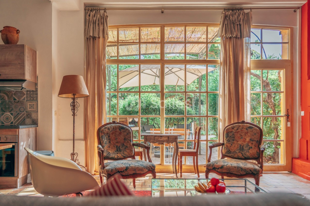 Salon lumineux avec fauteuils anciens, lampadaire et vue sur une terrasse ombragée par un parasol et un jardin verdoyant.
