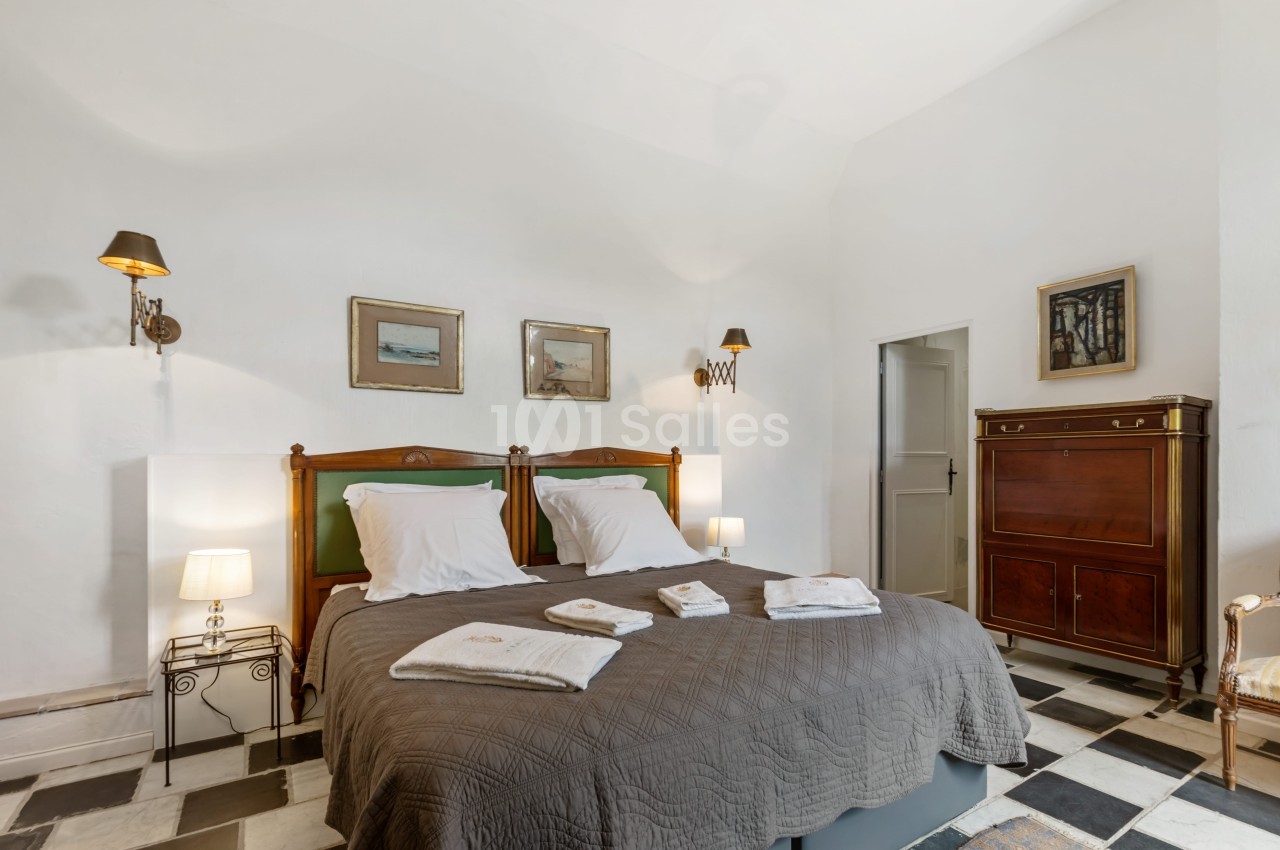 Chambre lumineuse avec lit double, linge de lit plié, mobilier ancien, murs blancs et sol en carrelage noir et blanc.