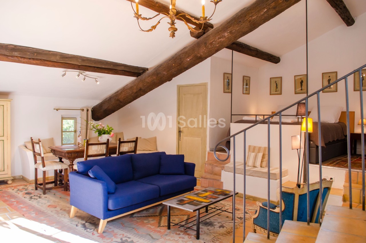 Salon lumineux avec canapé bleu, table à manger en bois, poutres apparentes et lit en mezzanine dans un intérieur chaleureux.