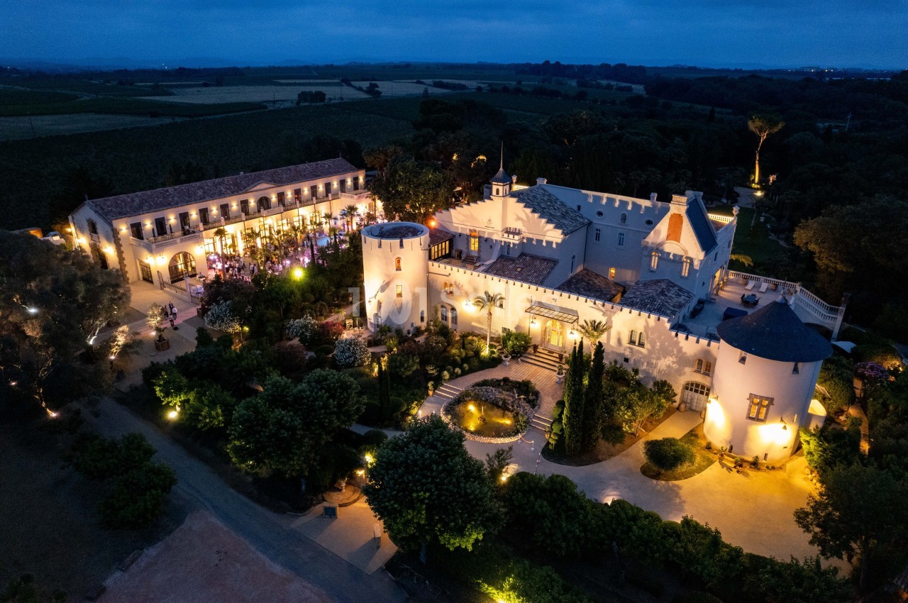 Vue aérienne d'un château éclairé de nuit, entouré de jardins et de bâtiments annexes dans un paysage rural.