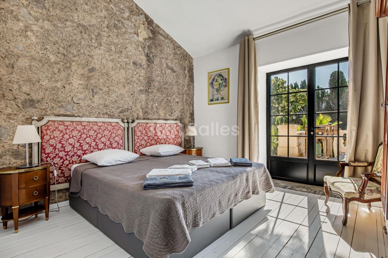 Chambre lumineuse avec lit double, tête de lit rouge, murs en pierre, grandes fenêtres et mobilier classique.