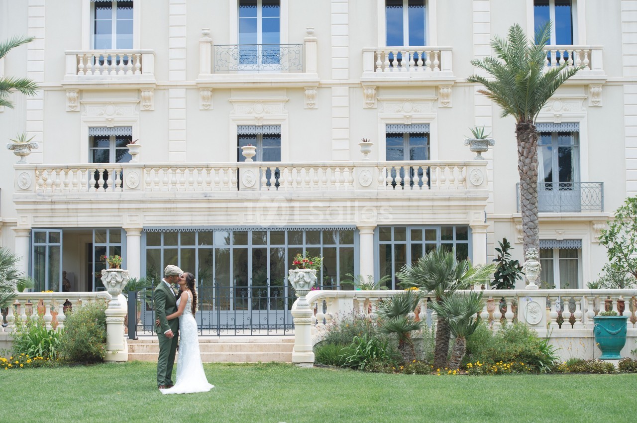 Un couple en tenue de mariage se tient devant un bâtiment élégant avec une façade beige et un jardin verdoyant.