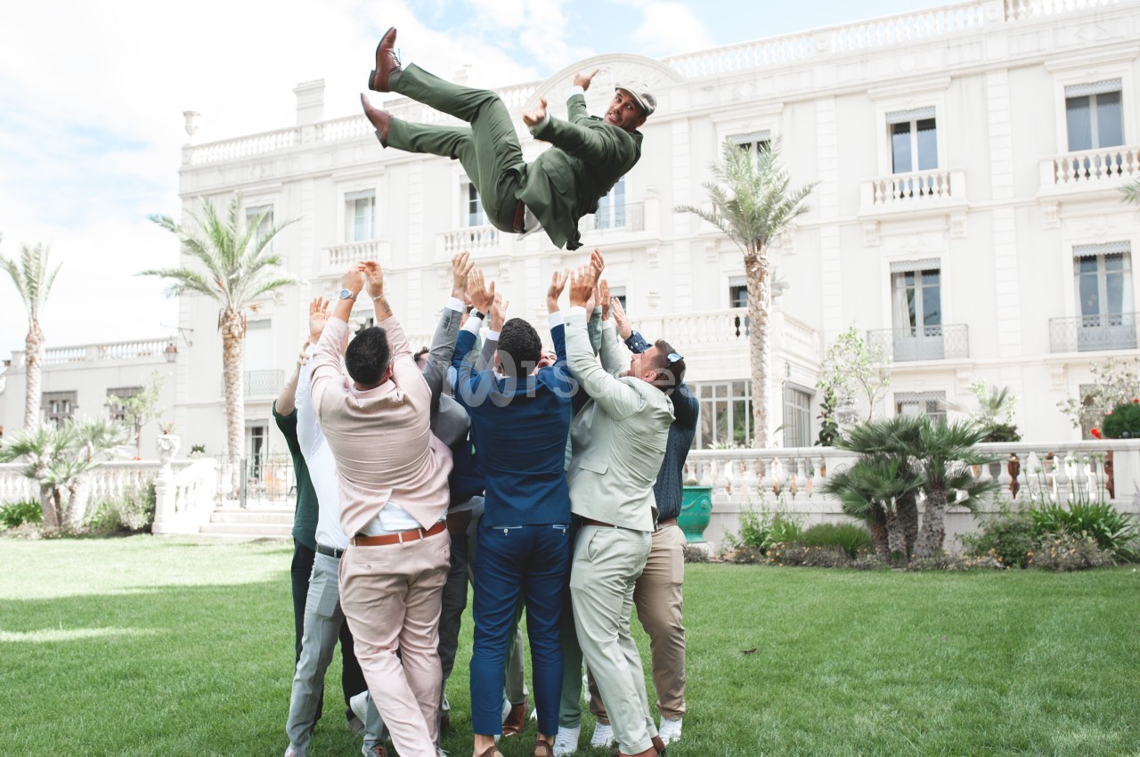 Un groupe d'hommes en tenue formelle lance un homme en costume vert en l'air devant un bâtiment élégant.