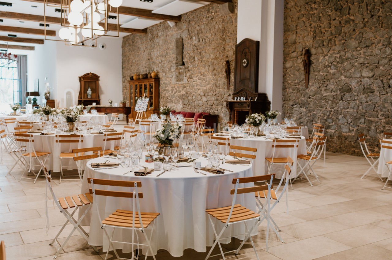Salle de réception décorée avec des tables rondes dressées, chaises en bois et murs en pierre apparente.