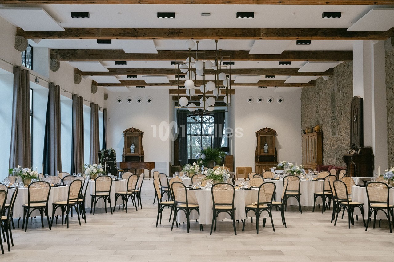 Salle de réception lumineuse avec tables rondes dressées, chaises en bois clair et poutres apparentes au plafond.