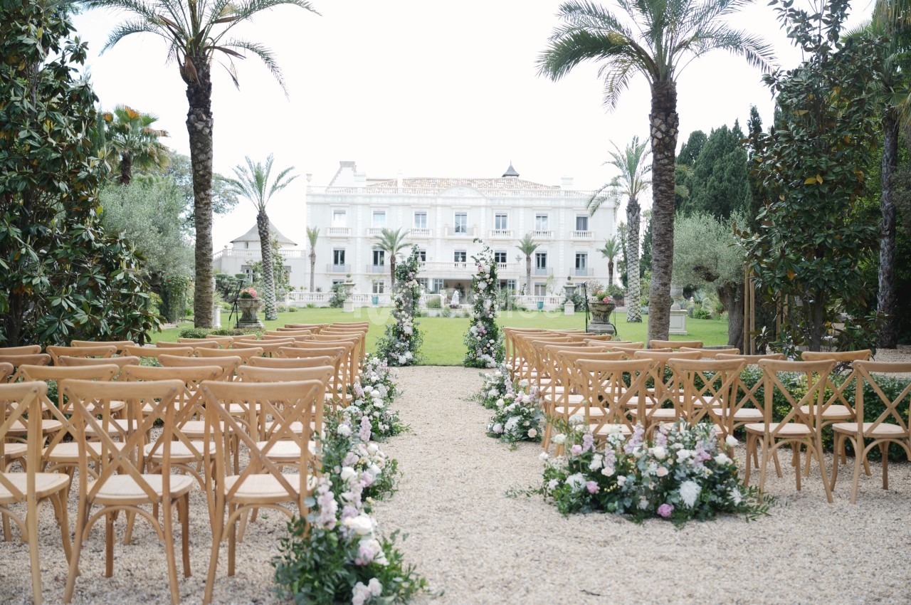 Allée centrale bordée de chaises en bois et de fleurs menant à un bâtiment blanc entouré de palmiers.