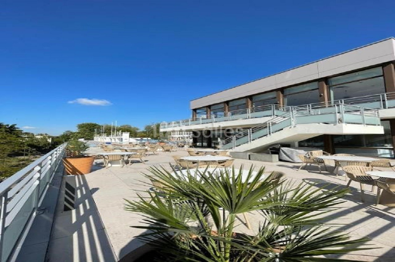 Terrasse ensoleillée avec tables et chaises, bordée de plantes, devant un bâtiment moderne sous un ciel bleu.