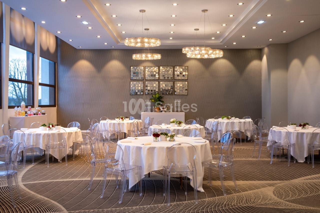 Salle de réception lumineuse avec tables rondes dressées, chaises transparentes et décorations modernes.