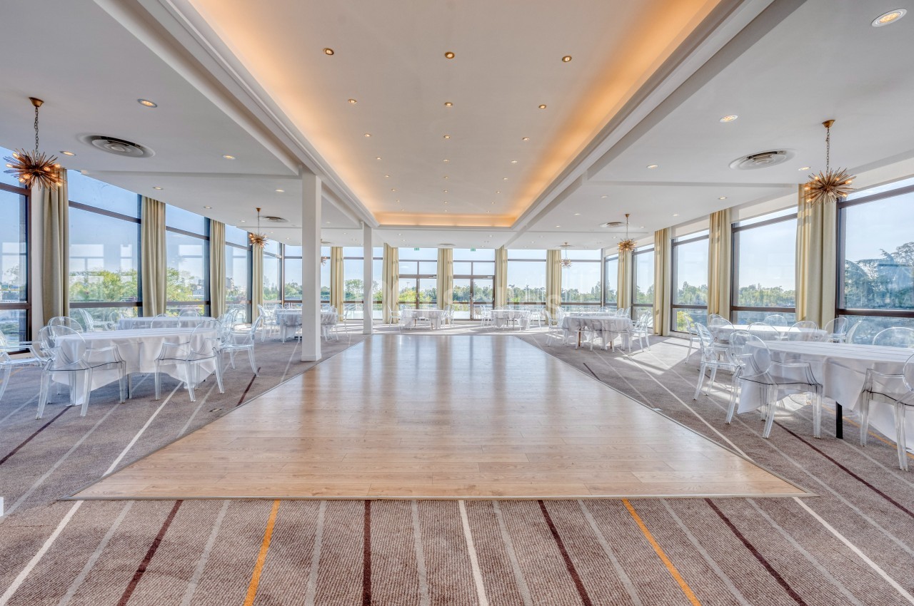 Salle lumineuse avec de grandes baies vitrées, tables rondes dressées et parquet central entouré de moquette.