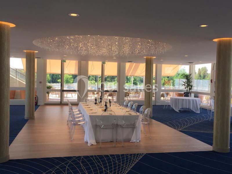 Salle lumineuse avec une grande table dressée, chaises transparentes, lustre moderne et baies vitrées donnant sur l…