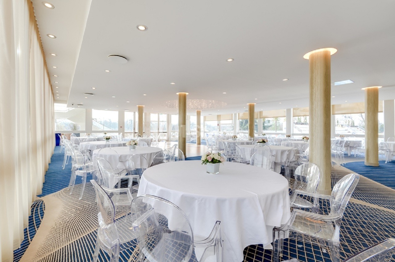 Salle lumineuse avec grandes baies vitrées, tables rondes dressées de nappes blanches et chaises transparentes.