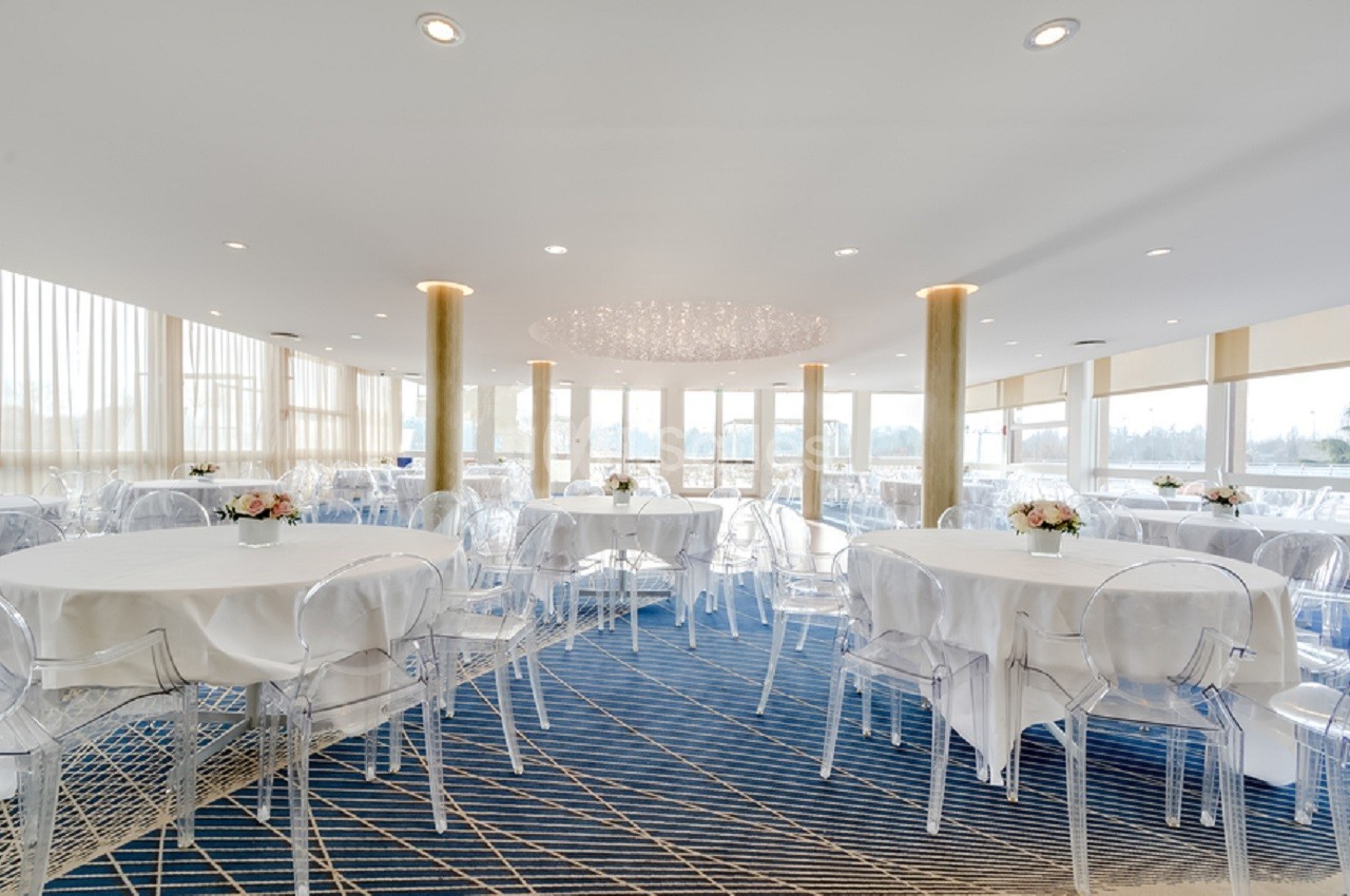 Salle lumineuse avec tables rondes dressées, chaises transparentes et grandes baies vitrées donnant sur l'extérieur.