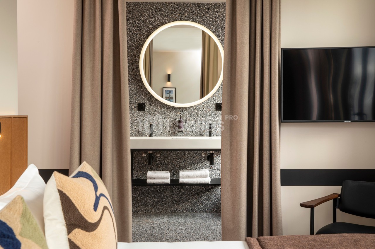 Vue d'une chambre d'hôtel avec un lit, un miroir rond éclairé et un lavabo en arrière-plan.