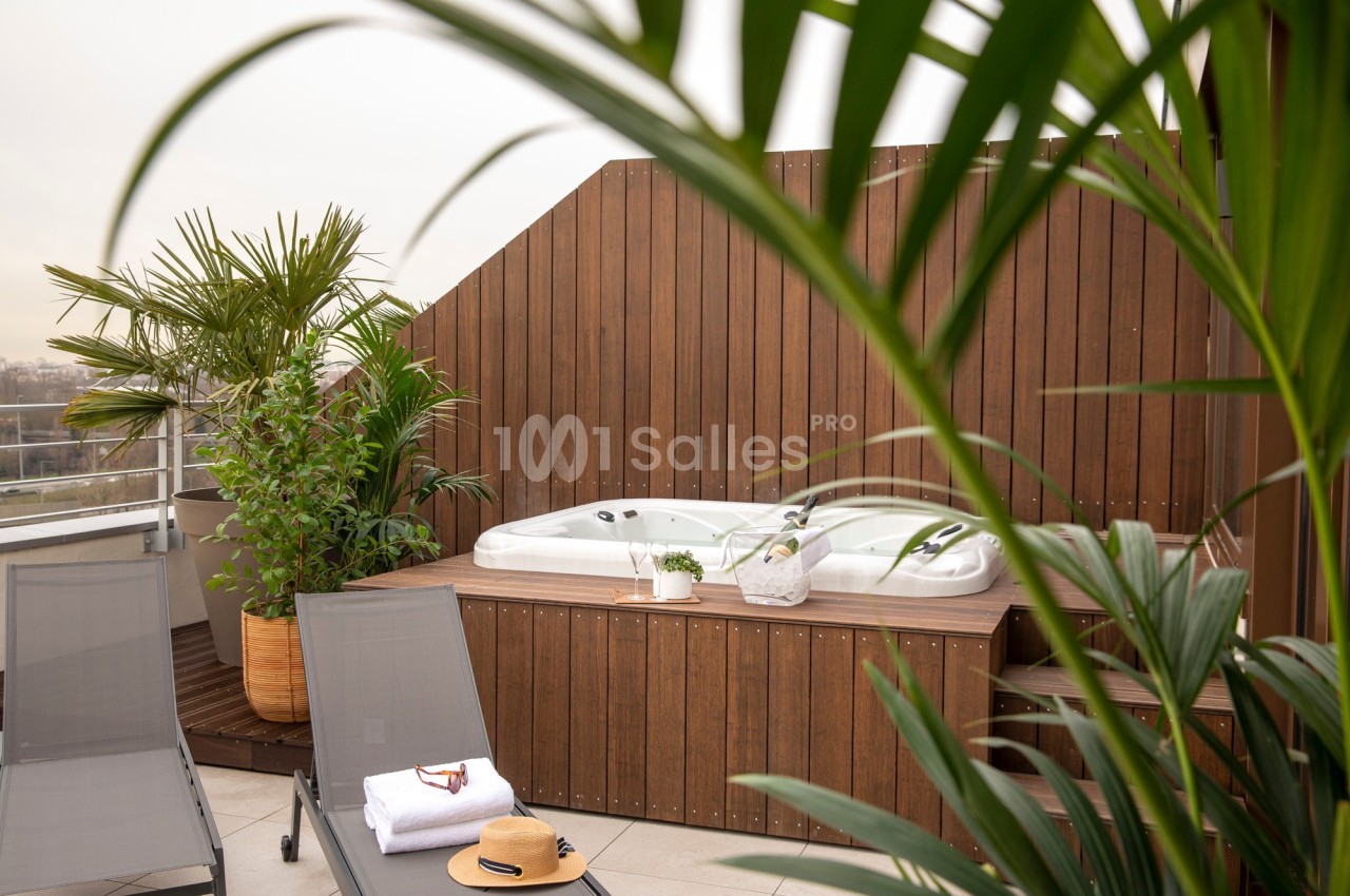 Jacuzzi extérieur entouré de plantes et de chaises longues sur une terrasse avec un panneau en bois.