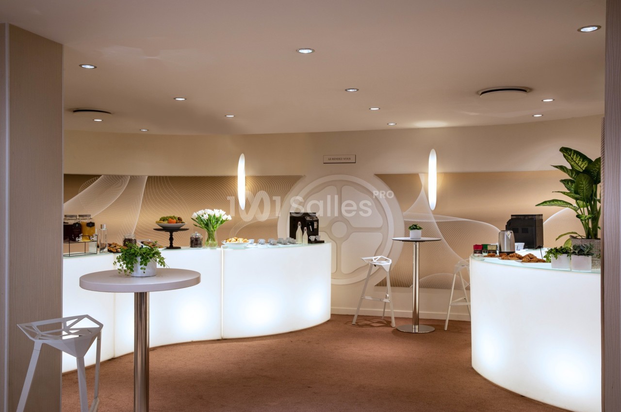 Espace lumineux avec tables hautes, plantes, machines à café et buffet disposés dans un cadre moderne et épuré.