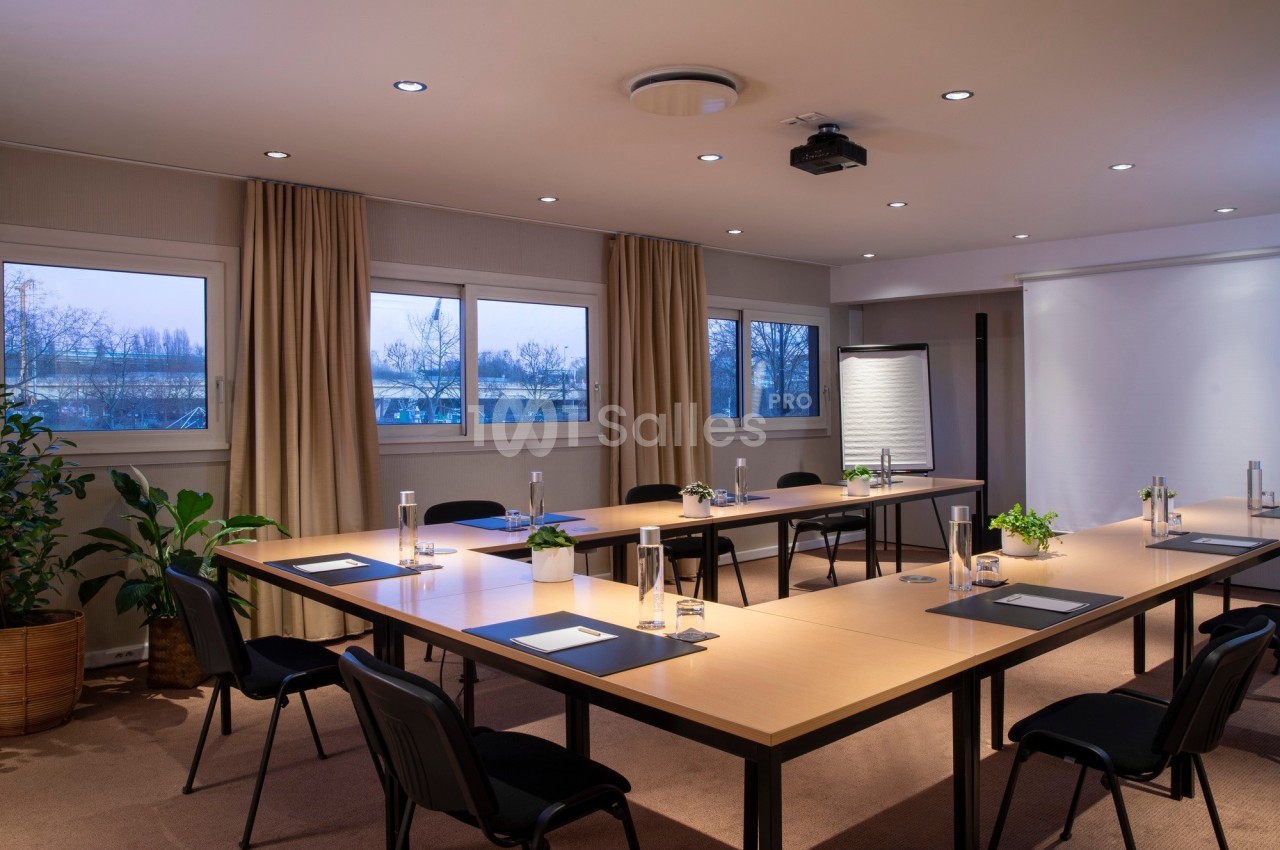 Salle de réunion lumineuse avec tables en U, chaises noires, plantes décoratives et matériel de présentation.