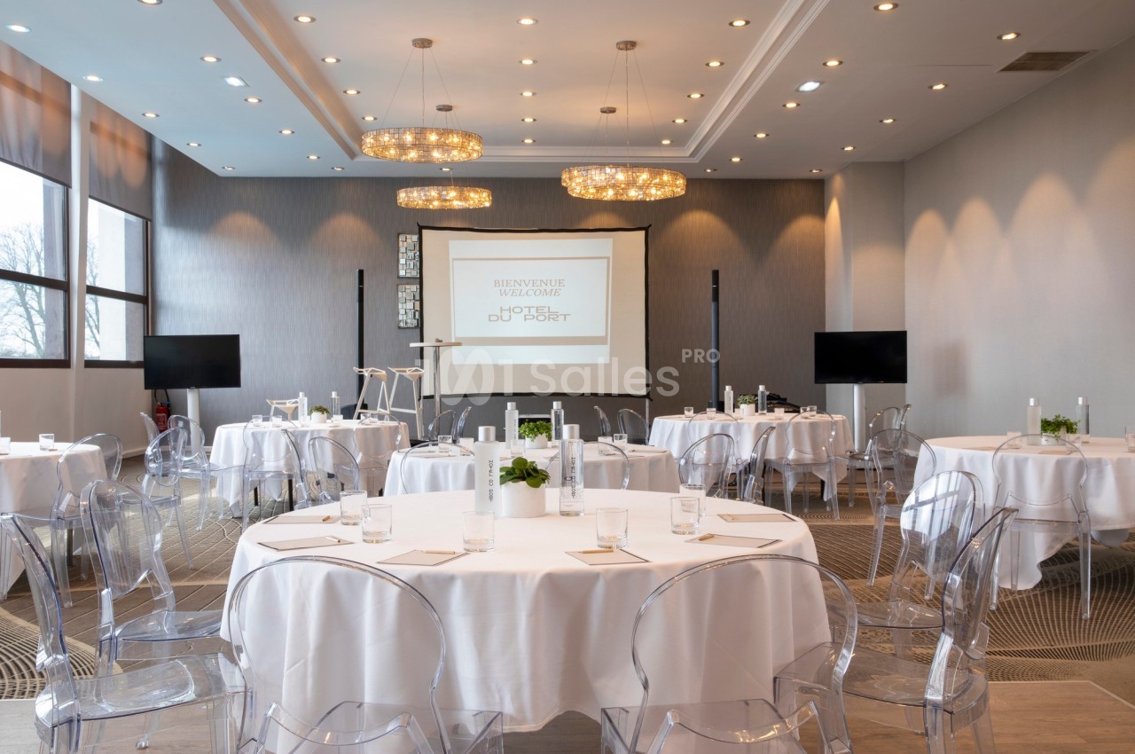 Salle de conférence lumineuse avec tables rondes dressées, chaises transparentes et écran de projection allumé.