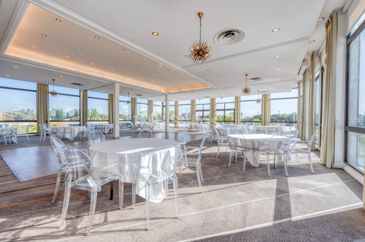 Salle lumineuse avec de grandes baies vitrées, tables rondes dressées de nappes blanches et chaises transparentes.
