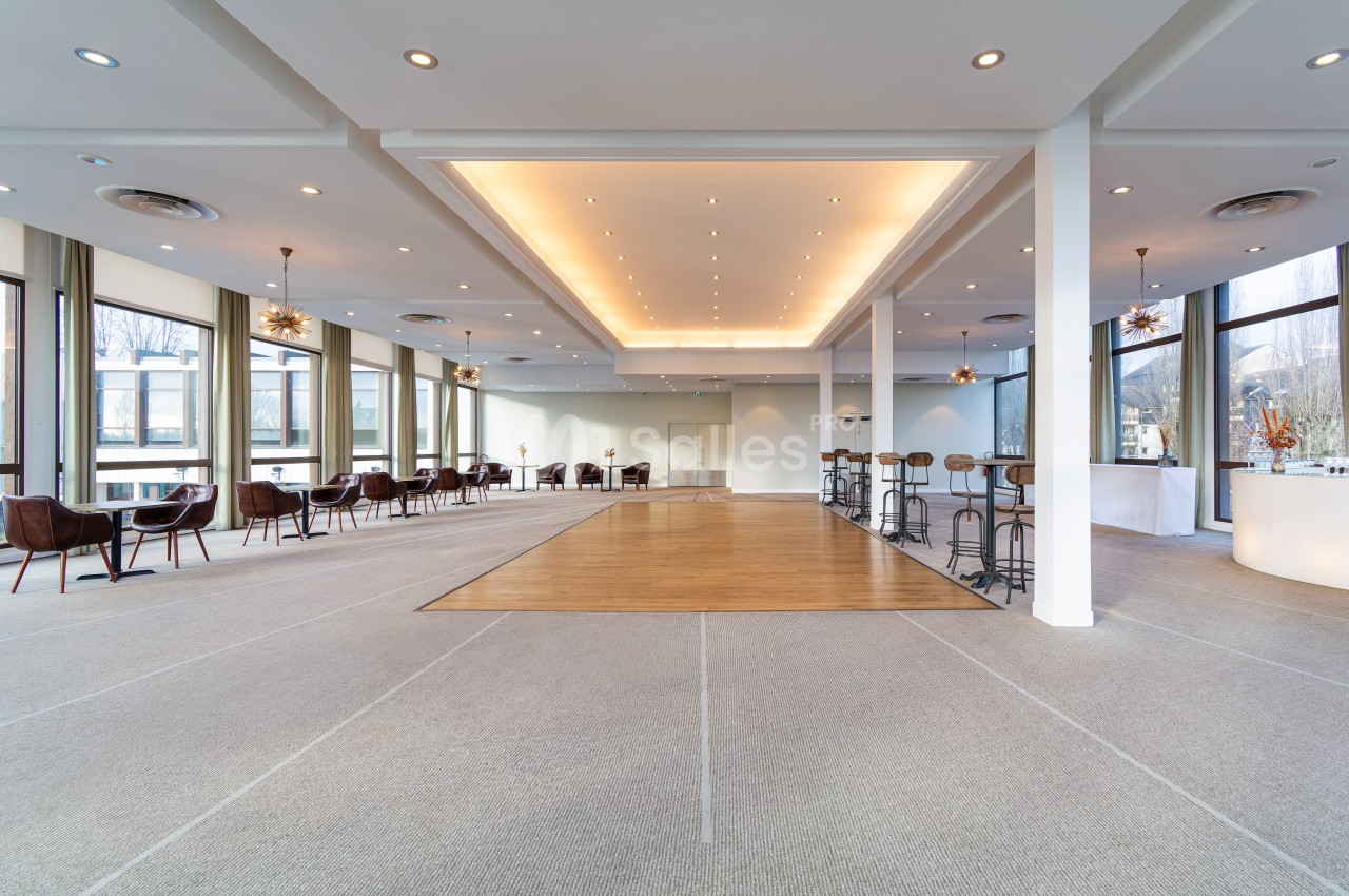 Salle lumineuse avec de grandes baies vitrées, un sol en moquette et une zone centrale en parquet, aménagée avec des tables…