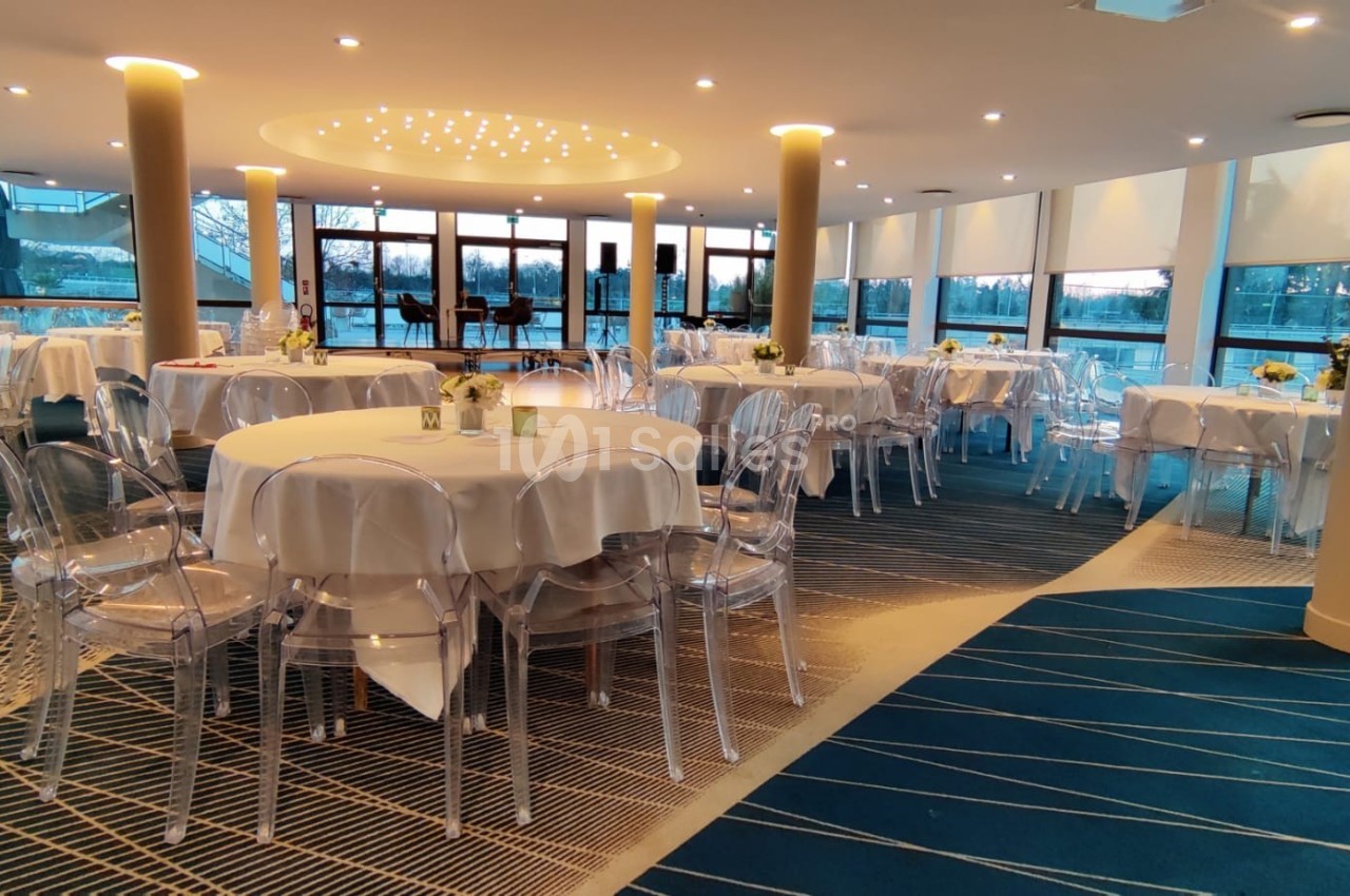 Salle lumineuse avec tables rondes dressées, chaises transparentes et grandes baies vitrées donnant sur l'extérieur.